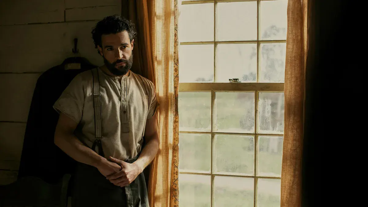 Christopher Abbott en Adam Trask, barbu et en bretelles, adossé près d'une fenêtre dans la série À l'est d'Éden