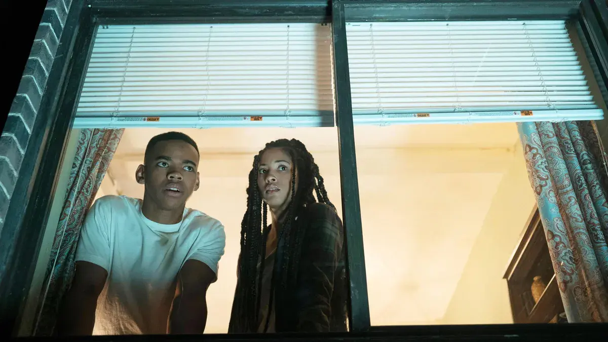 Joivan Wade et Lex Scott Davis guettent la rue depuis une fenêtre de Staten Island, dans American Nightmare 4 : Les Origines