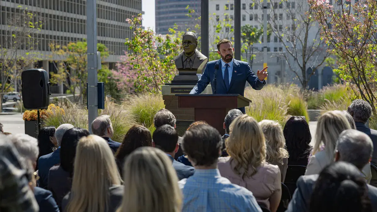 Ben Affleck en costume bleu, debout derrière un pupitre, s'adresse à une foule assise en plein air dans le film Animals sur Netflix