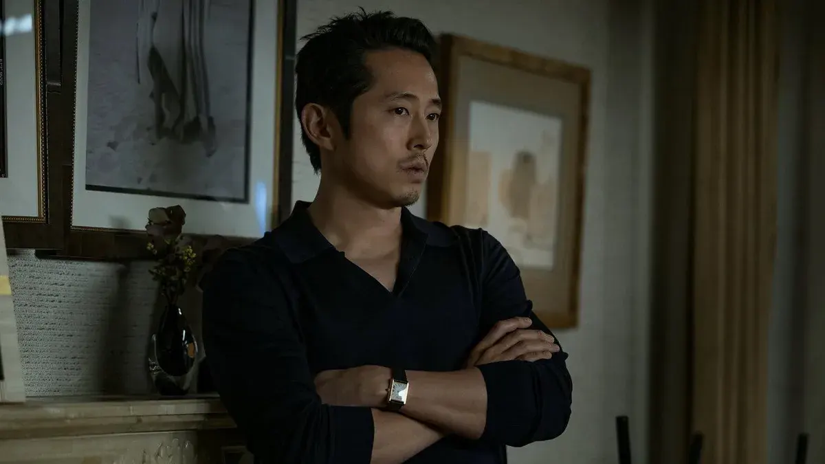 Steven Yeun, bras croisés et pull noir, adossé à une cheminée dans un salon cossu, dans le film Animals de Ben Affleck