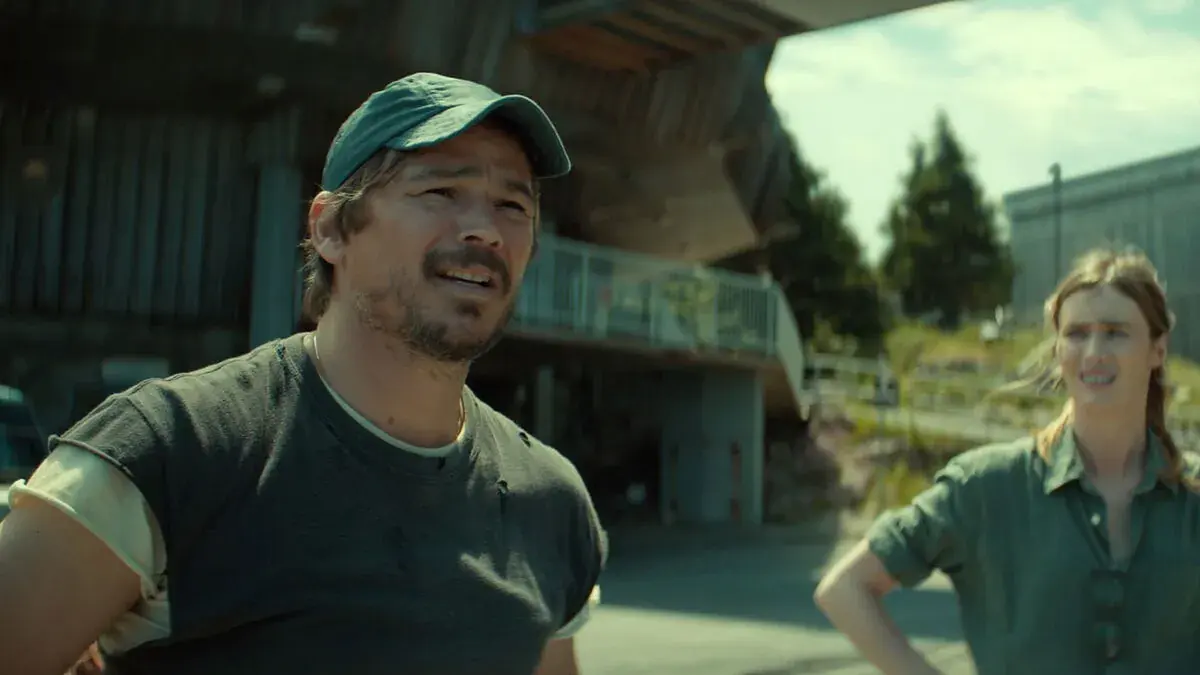 Josh Hartnett, casquette verte et t-shirt usé, fronce les sourcils au soleil devant un bâtiment de béton, Mackenzie Davis en chemise verte derrière lui dans Below