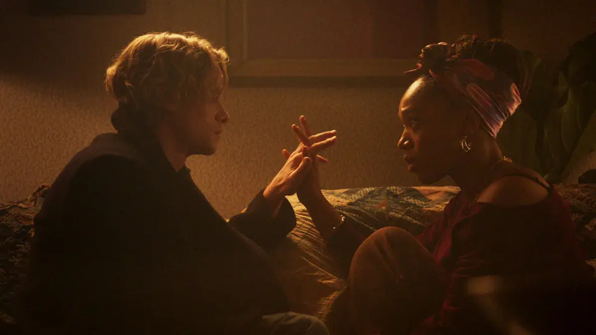 Tom Rhys Harries en Matt Hagen et Naomi Ackie en Caitlyn Corr, assis face à face, les mains jointes, dans une chambre éclairée d'une lumière ambrée, dans Clayface