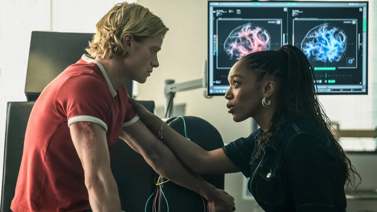 Tom Rhys Harries en Matt Hagen, polo rouge et bras écorché, face à Naomi Ackie dans un laboratoire devant un écran d'imagerie cérébrale, dans Clayface