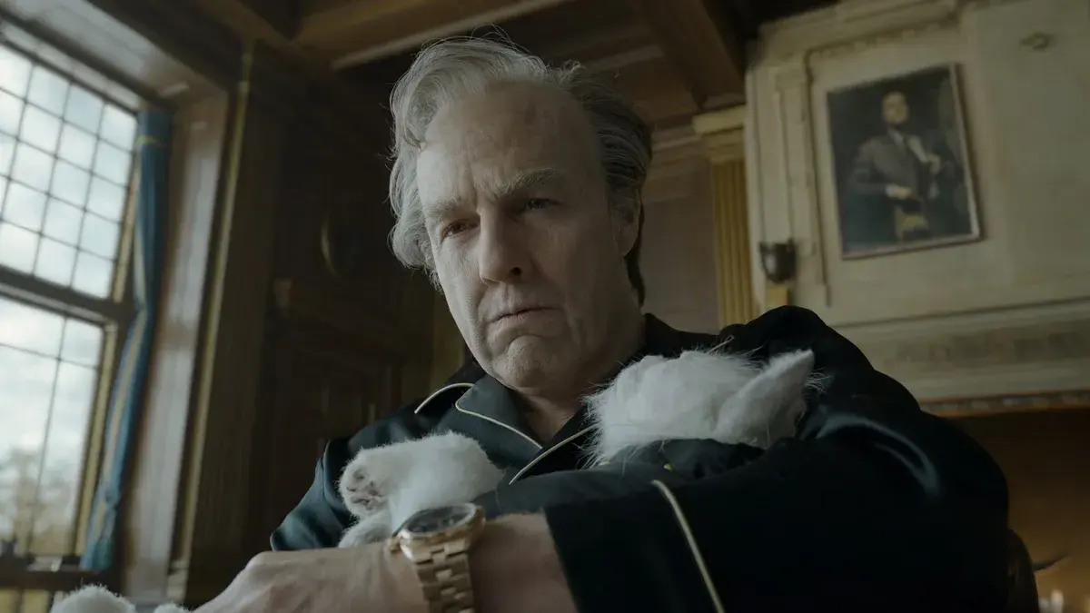 Tom Cruise, vieilli, en pyjama de soie noire et montre en or, serre un chat blanc contre lui dans un bureau lambrissé de Digger