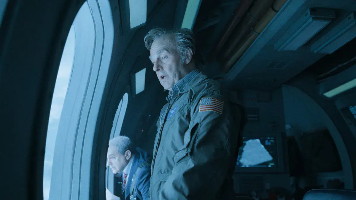 Tom Cruise, cheveux gris et combinaison de vol frappée du drapeau américain, devant le hublot d'un appareil dans Digger d'Alejandro G. Iñárritu