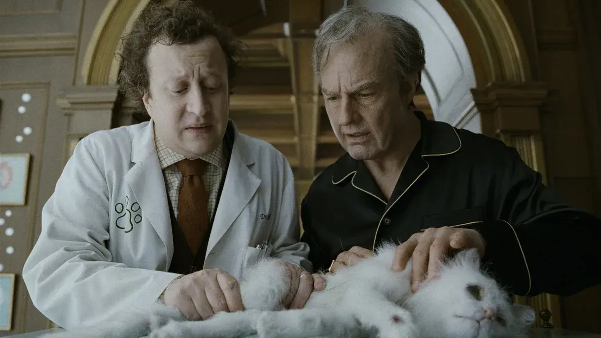 Tom Cruise, vieilli, en pyjama de soie noire, penché avec un vétérinaire sur un chat blanc allongé dans Digger d'Alejandro G. Iñárritu