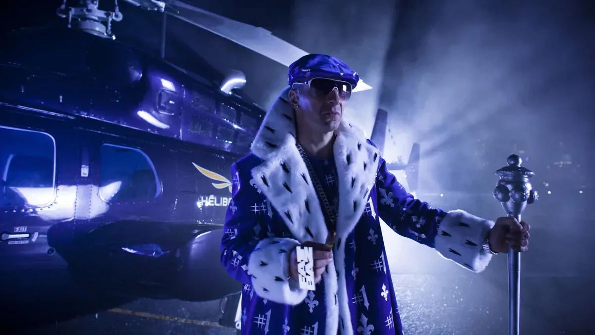 Michaël Youn en Fatal Bazooka, lunettes noires, casquette et manteau bleu à fleurs de lys, un sceptre à la main devant un hélicoptère dans Fatal (2010)