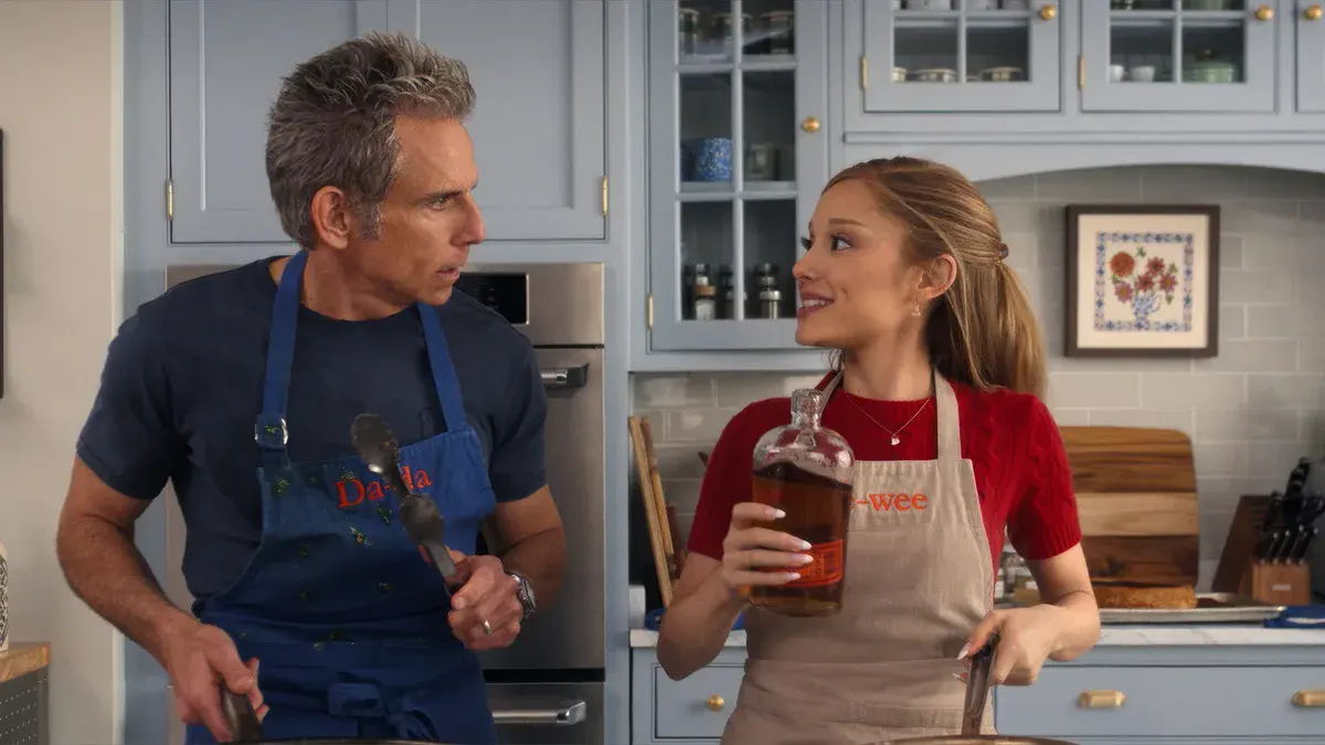 Ben Stiller en tablier et Ariana Grande, une bouteille à la main, dans une cuisine dans Mon beau-père, ma belle-fille et moi