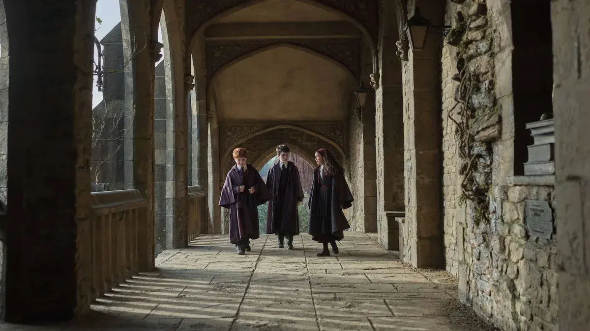 Ron, Harry et Hermione en robes de Poudlard marchent sous les arches d'un cloître dans la série Harry Potter de HBO
