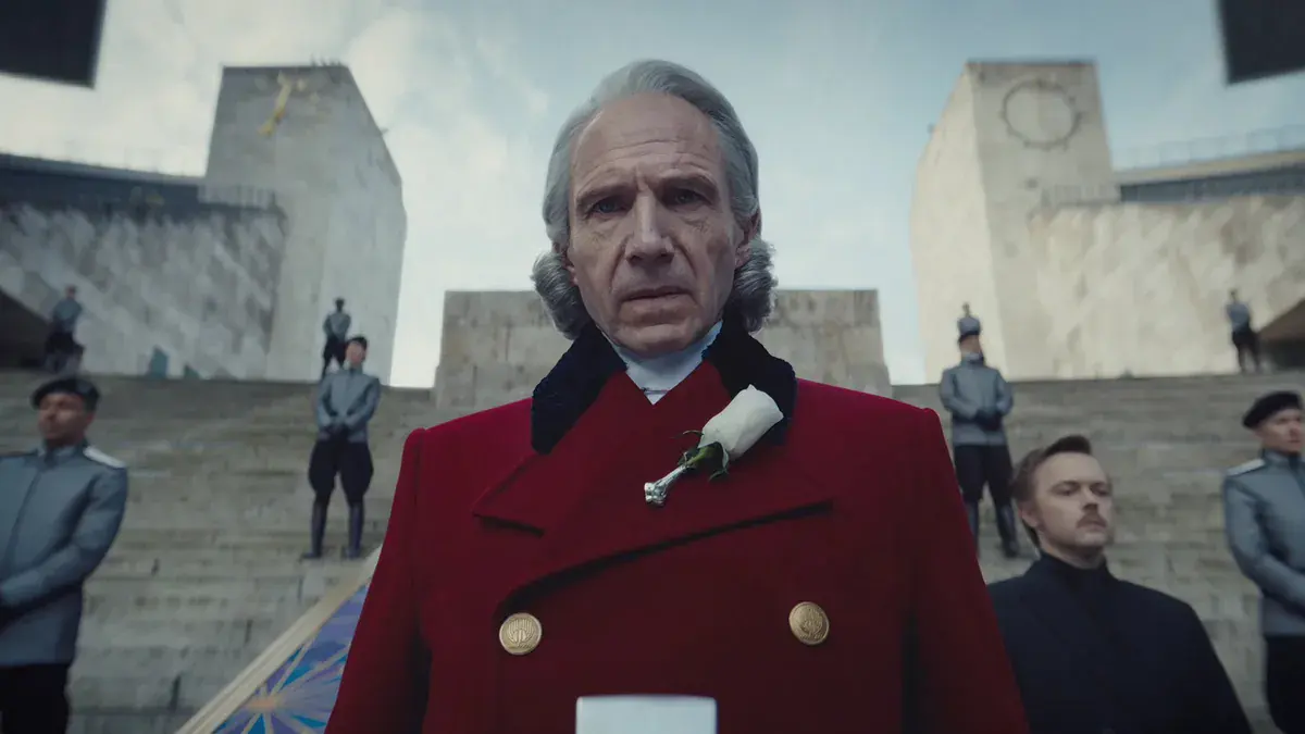 Ralph Fiennes en président Coriolanus Snow, manteau rouge et rose blanche à la boutonnière, devant un escalier gardé par des Pacificateurs dans Hunger Games : Lever de soleil sur la moisson