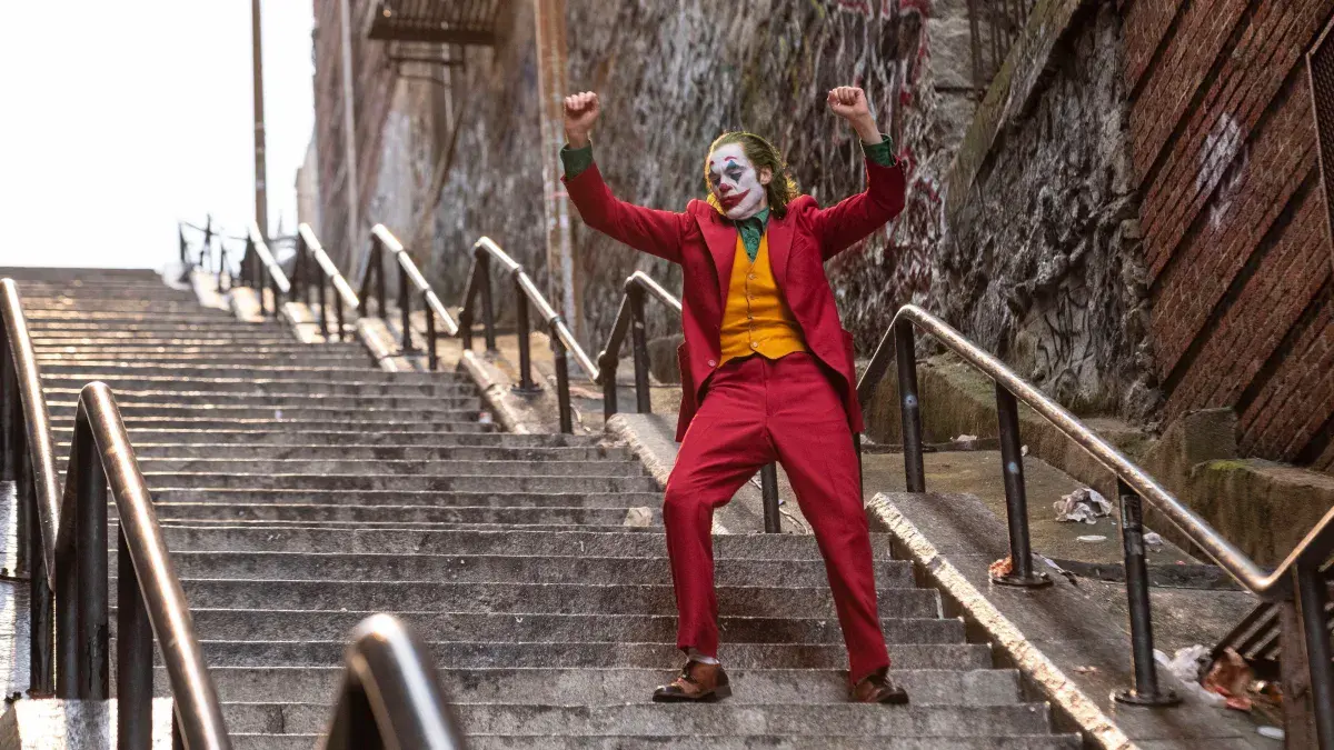 Joaquin Phoenix en Joker, costume rouge et maquillage de clown, danse les bras levés dans un escalier du Bronx