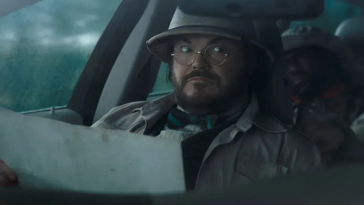 Jack Black, chapeau colonial et lunettes rondes, lit une carte à l'avant d'une voiture sous la pluie, Kevin Hart sur la banquette arrière, dans Jumanji : Bienvenue dans notre monde