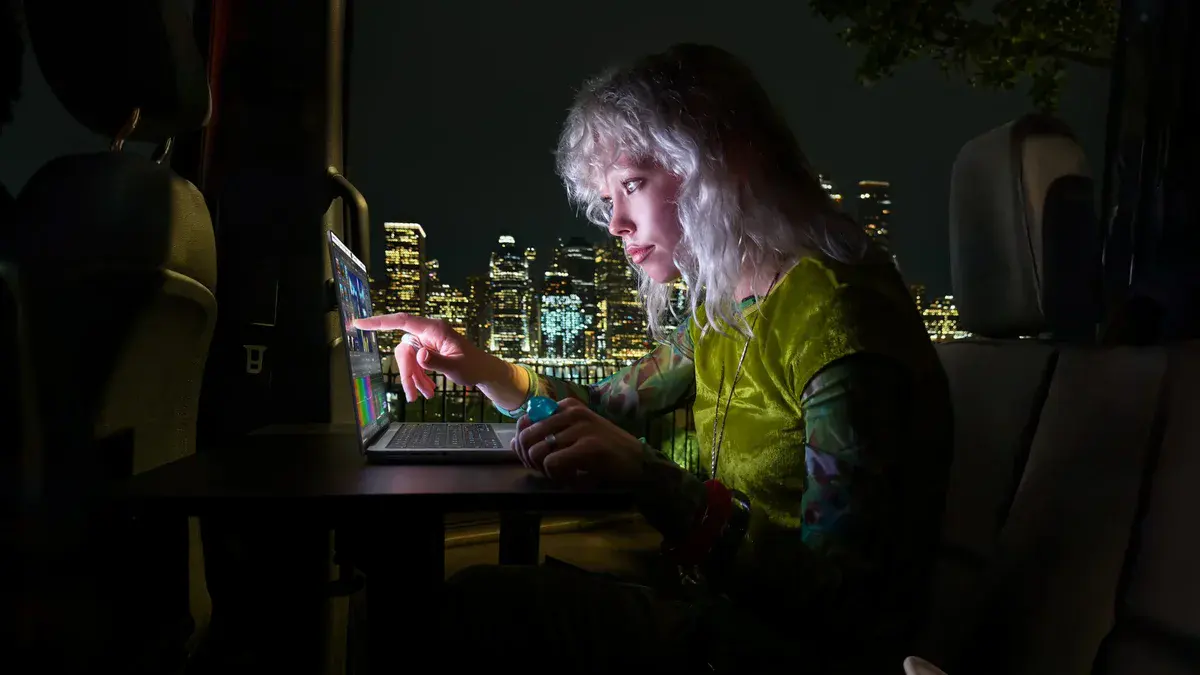 Une femme aux cheveux blonds touche l'écran de son MacBook Pro PROLED XDR, le soir, devant les lumières d'une ville