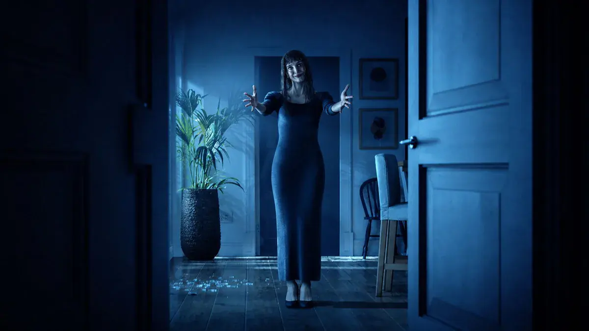 L'Autre Maman, jouée par Jessica Chastain, bras tendus et sourire figé au bout d'un couloir plongé dans la lumière bleue dans Malfaisante