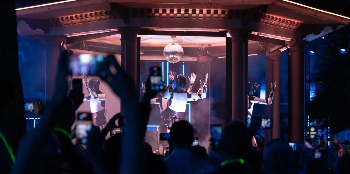Des robots Optimus dansent sous un kiosque éclairé devant un public qui filme avec des téléphones, lors de la soirée We, Robot organisée par Tesla en octobre 2024