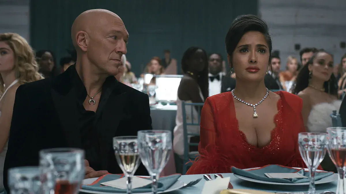 Vincent Cassel, crâne rasé et veste noire, assis à côté de Salma Hayek en robe rouge et collier de diamants, à une table de gala dans le film Sacrifice