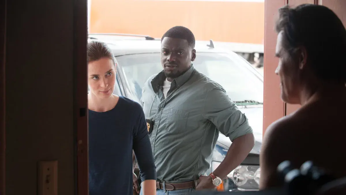 Emily Blunt en agent du FBI Kate Macer, à côté de Daniel Kaluuya, sur le seuil d'une porte face à Josh Brolin vu de dos, dans Sicario de Denis Villeneuve