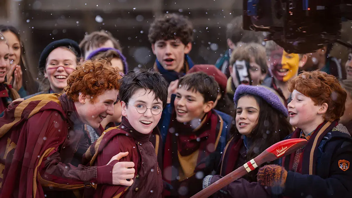 Harry, en robe de Gryffondor, entouré des Weasley et d'élèves de Poudlard sous la neige, dans la série Harry Potter de HBO