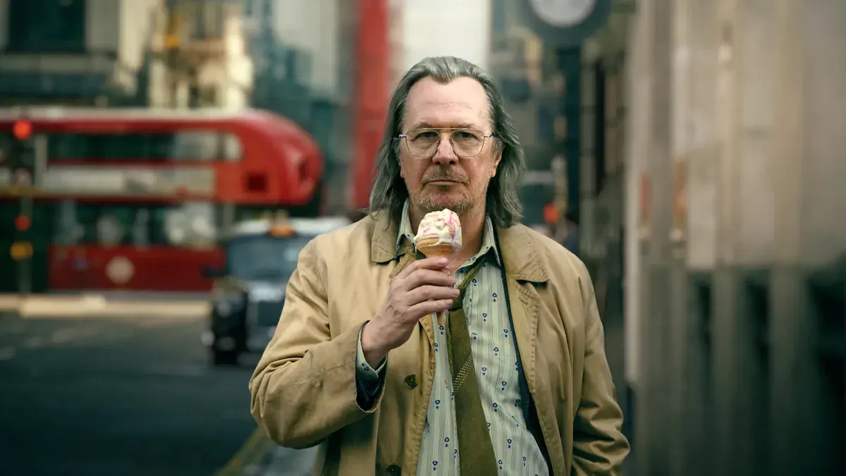Gary Oldman en Jackson Lamb, imperméable beige et cornet de glace à la main, dans une rue de Londres, dans Slow Horses