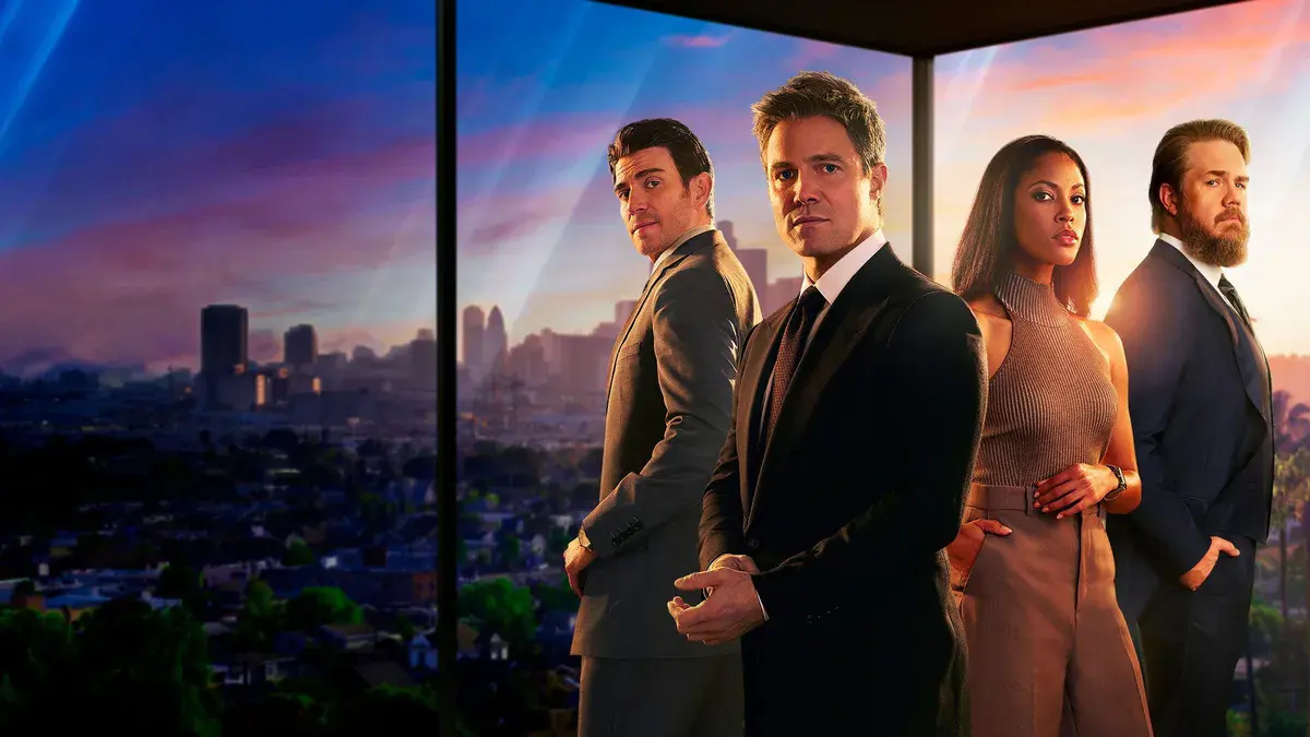 Suits LA : Stephen Amell en Ted Black, costume sombre, entouré des avocats de son cabinet devant une baie vitrée qui domine Los Angeles au coucher du soleil