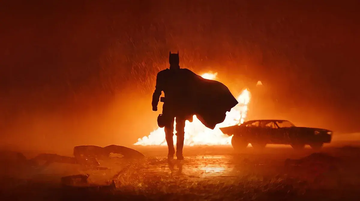 Batman, cape au vent, marche sous la pluie devant une voiture en flammes dans The Batman de Matt Reeves (2022)