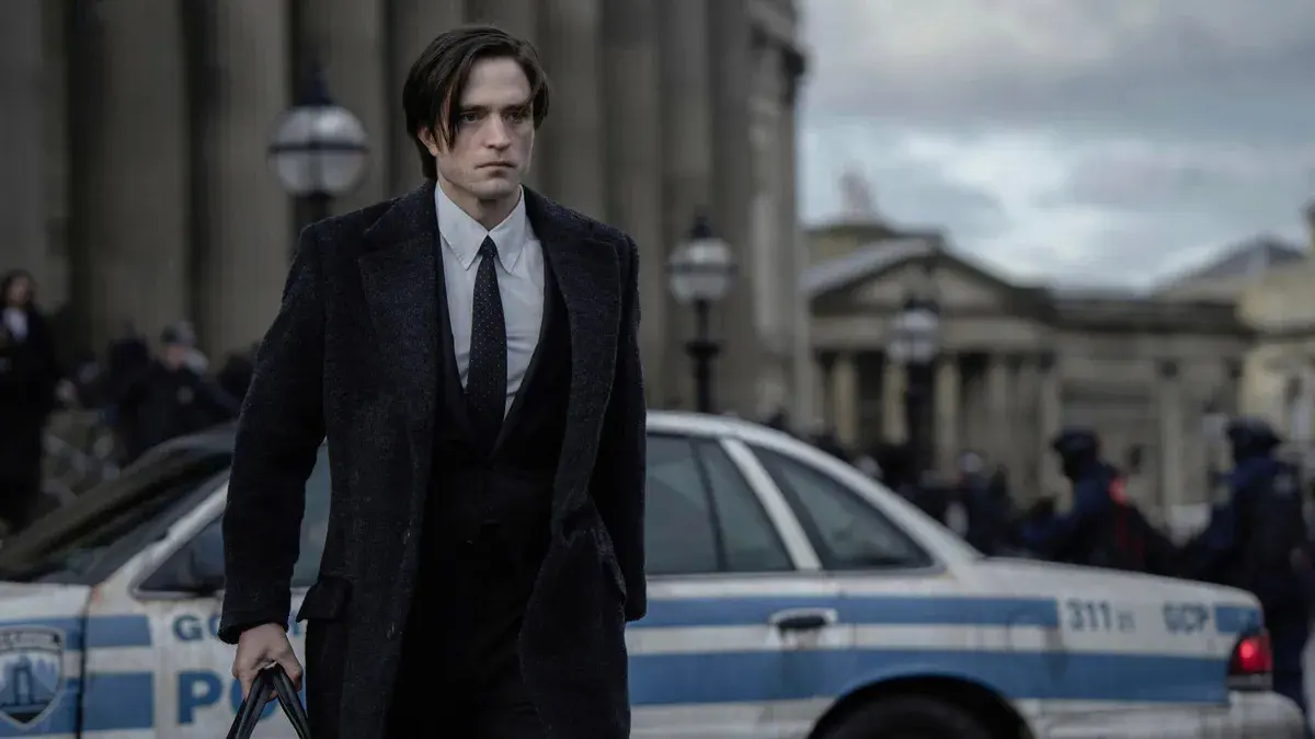 Robert Pattinson en Bruce Wayne, manteau noir et sac à la main, devant une voiture de la police de Gotham dans The Batman (2022)