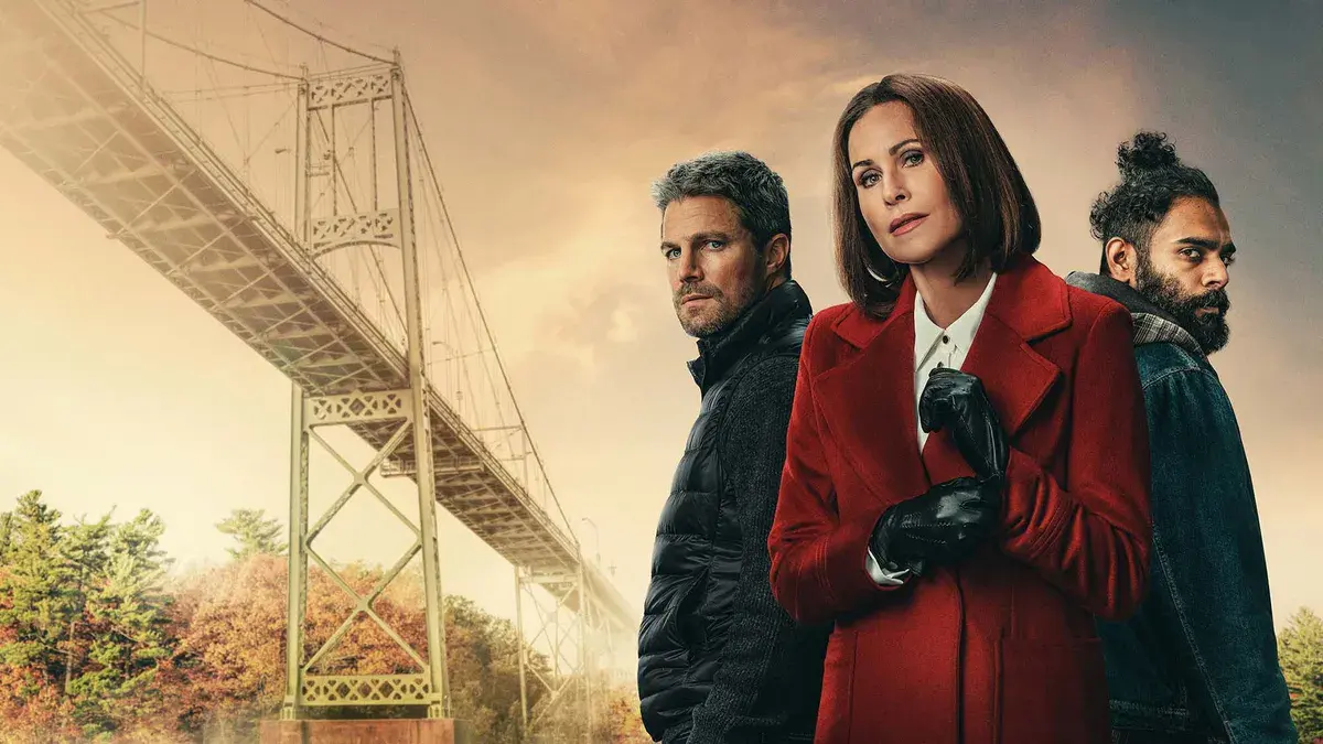 The Borderline : Stephen Amell, Minnie Driver en manteau rouge et Hamza Haq sous un pont suspendu des Mille-Îles, dans le ciel orangé de l'automne