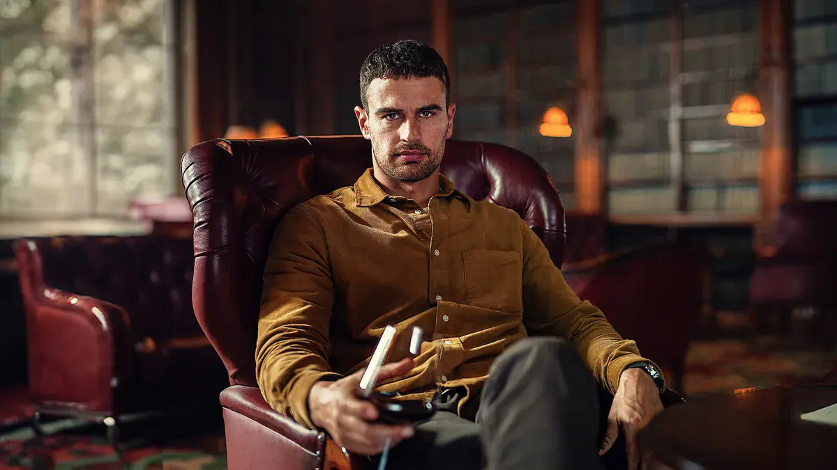Theo James en Eddie Horniman, assis dans un fauteuil en cuir, un verre à la main, dans une bibliothèque de The Gentlemen sur Netflix