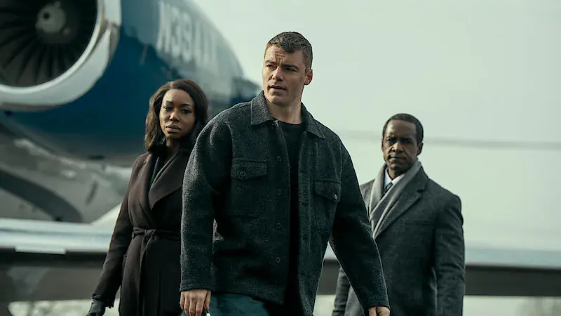 Gabriel Basso, en veste grise, marche devant un jet privé, suivi de deux personnages en manteau, dans The Night Agent