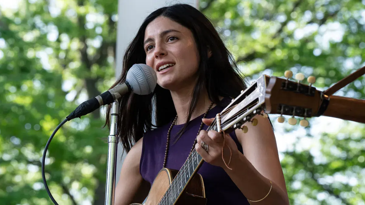 Monica Barbaro en Joan Baez chante à la guitare sur une scène en plein air dans Un parfait inconnu