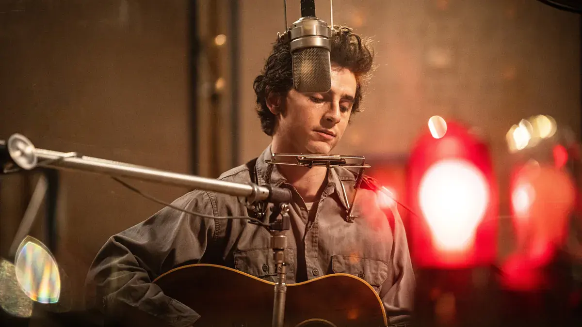 Timothée Chalamet en Bob Dylan, guitare acoustique et harmonica, chante devant un micro de studio dans Un parfait inconnu
