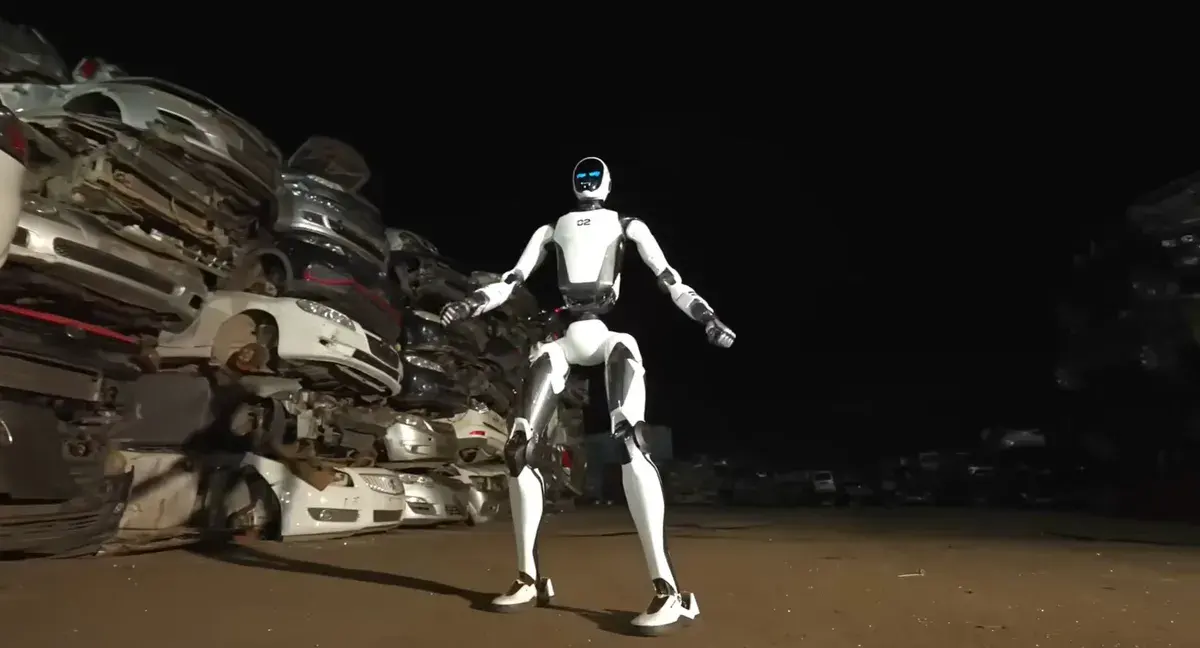 Le robot humanoïde Unitree H2-X, blanc à visage lumineux bleu, debout de nuit devant une pile de voitures dans une casse automobile