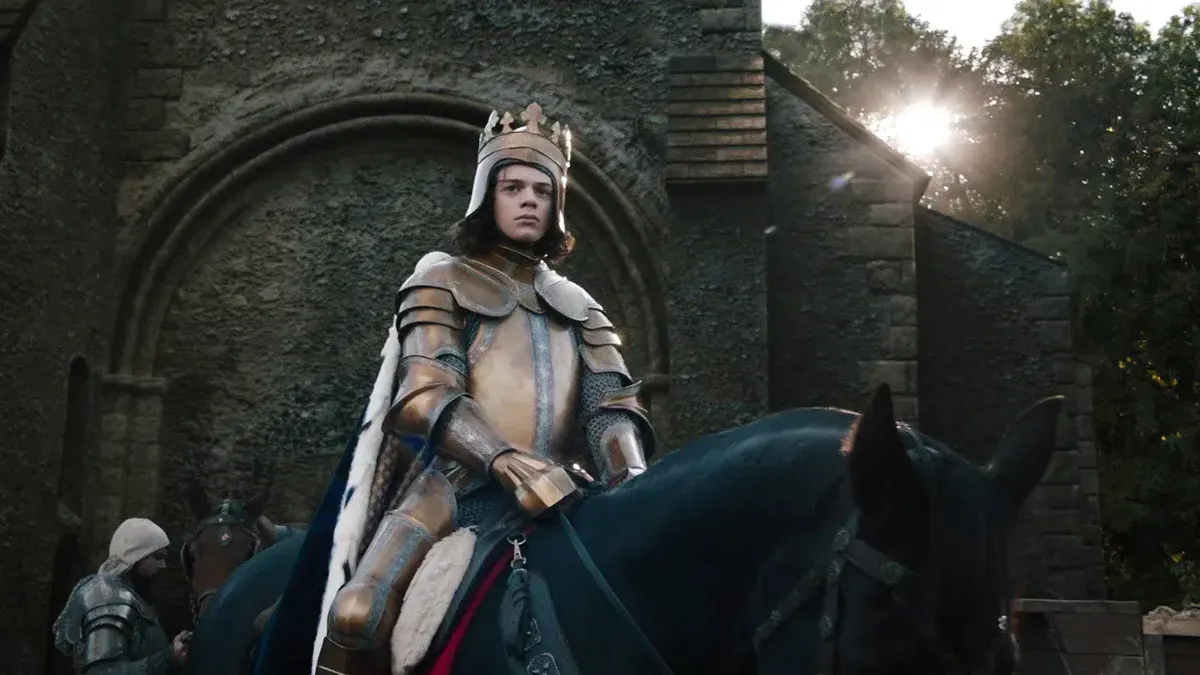 Woody Norman en Richard II, couronné et en armure dorée, à cheval devant une église dans The Uprising