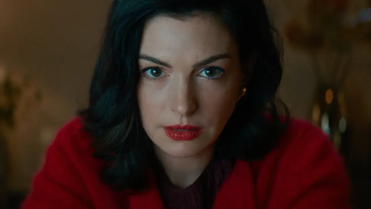 Anne Hathaway, rouge à lèvres et manteau rouge, fixe la caméra dans le rôle de Verity Crawford dans Verity