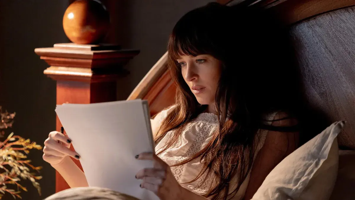 Dakota Johnson, allongée dans un lit en bois, lit les feuillets du manuscrit de Verity Crawford dans Verity