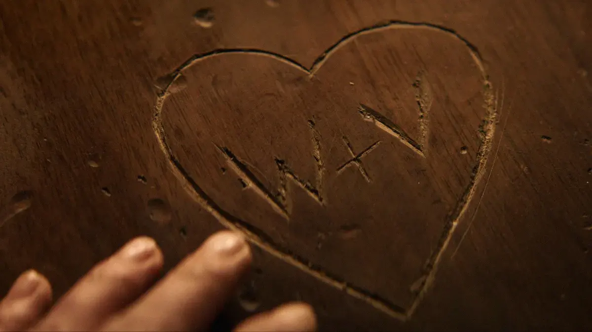 Un cœur gravé dans une table en bois avec les initiales W et V, effleuré par une main, dans une image de la série VisionQuest
