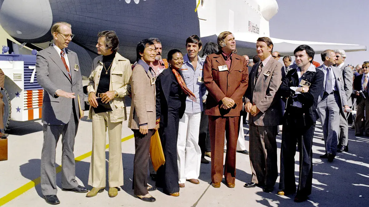 Walter Koenig, à droite en veste sombre, avec Leonard Nimoy, George Takei, Nichelle Nichols, DeForest Kelley, James Doohan et Gene Roddenberry devant la navette spatiale Enterprise de la NASA en 1976